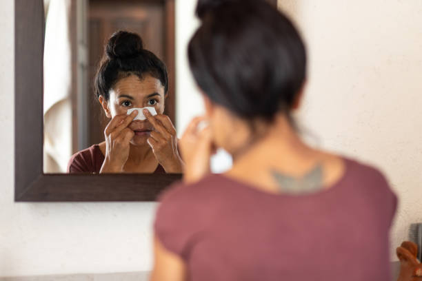 Woman in mirror applying a nasal strip to her nose for skincare or breathing.