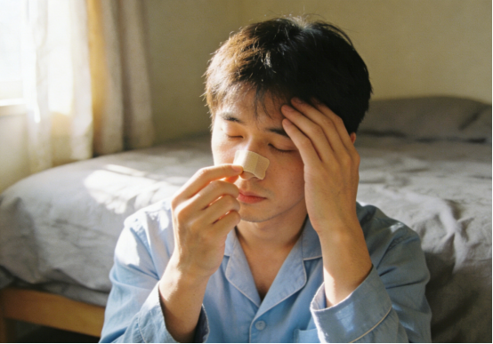 Man applying nasal strips on his nose while sitting on floor, feeling unwell.