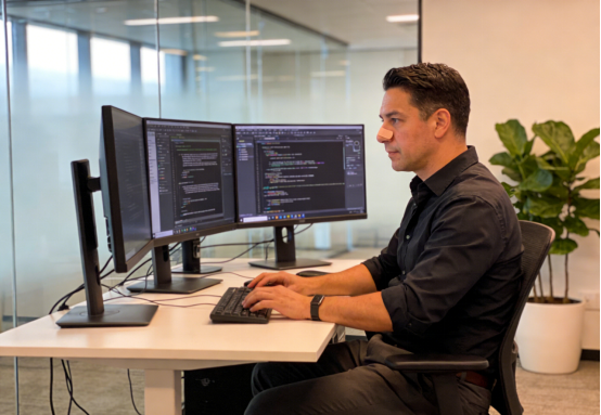 Man works intently at computer monitors, often assisted by nasal strips for focus.